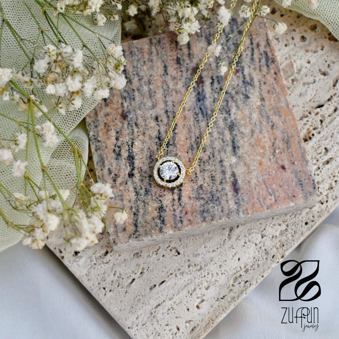 Gold necklace with a diamond pendant on a stone surface with flowers, Zulfur brand visible